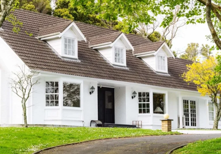 White two-story house with a brown tiled roof, dormer windows, green lawn, and a curved asphalt driveway.